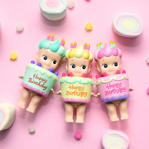 Three Sonny Angel birthday cake dolls with pastel hair and candles on a pink background with marshmallows