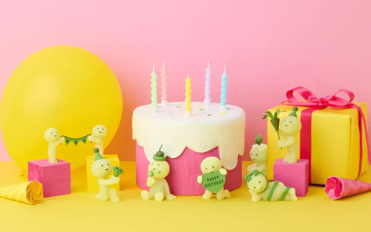Smiski glow-in-the-dark figures celebrating with a pink birthday cake, candles, yellow balloon, and gift box