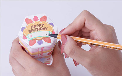 Person writing on a colorful Happy Birthday gift box with fruits, featuring Sonny Angel and Pop Mart style